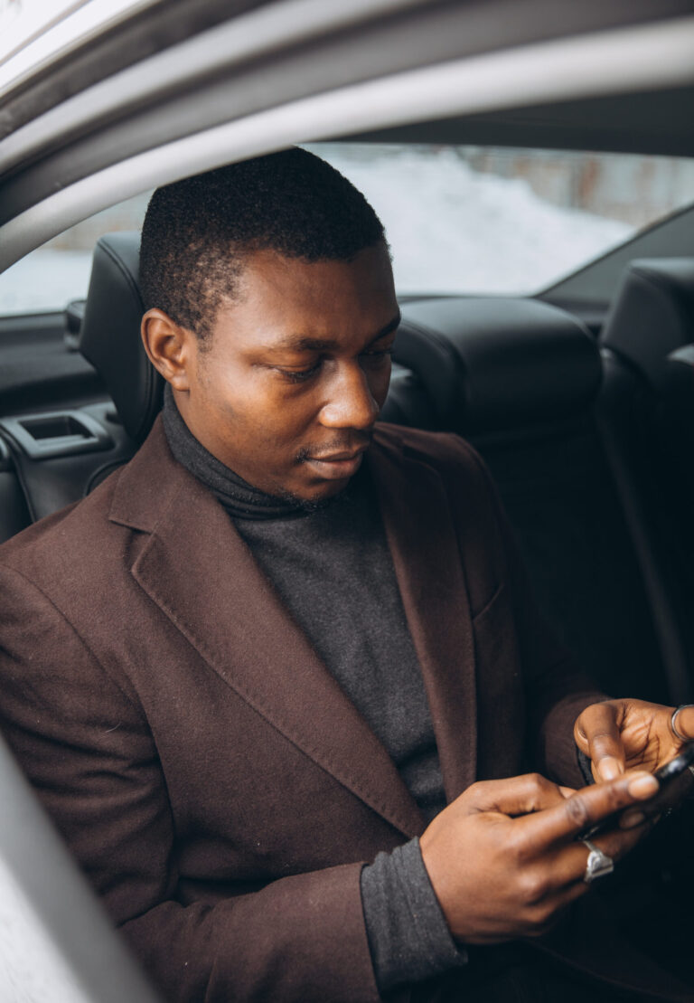 Young African American man sitting in a car's back seat, using a smartphone. Capturing business communication on the go